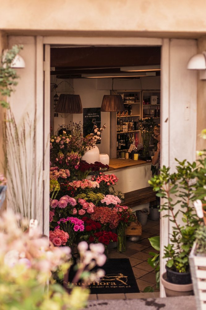 Floristería abierta con muchas flores. Dentro se ve a la dependienta y una clienta comprando.